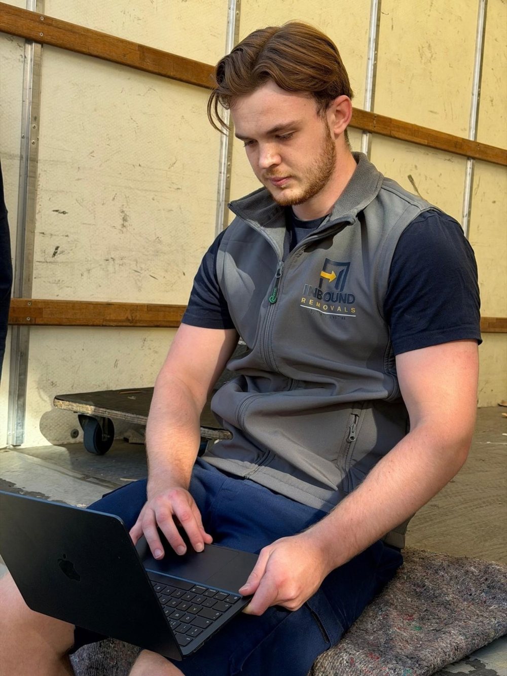 Harry McCarron on the back of a removals van with his laptop, wearing an Inbound Removals gilet